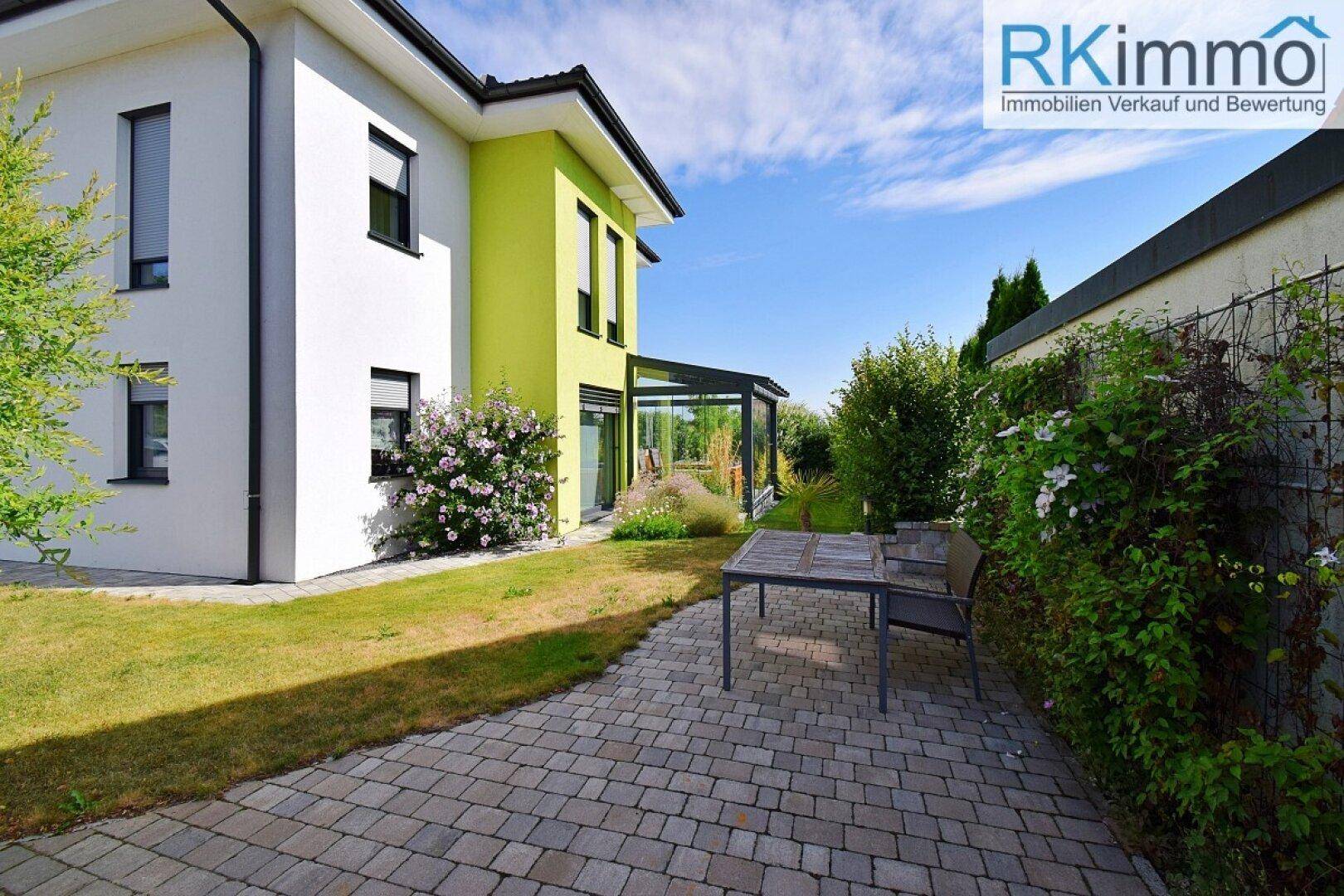 Seitlicher Blick auf das Haus mit gepflasterter Terrasse, Gartenmöbeln und üppiger Bepflanzung.