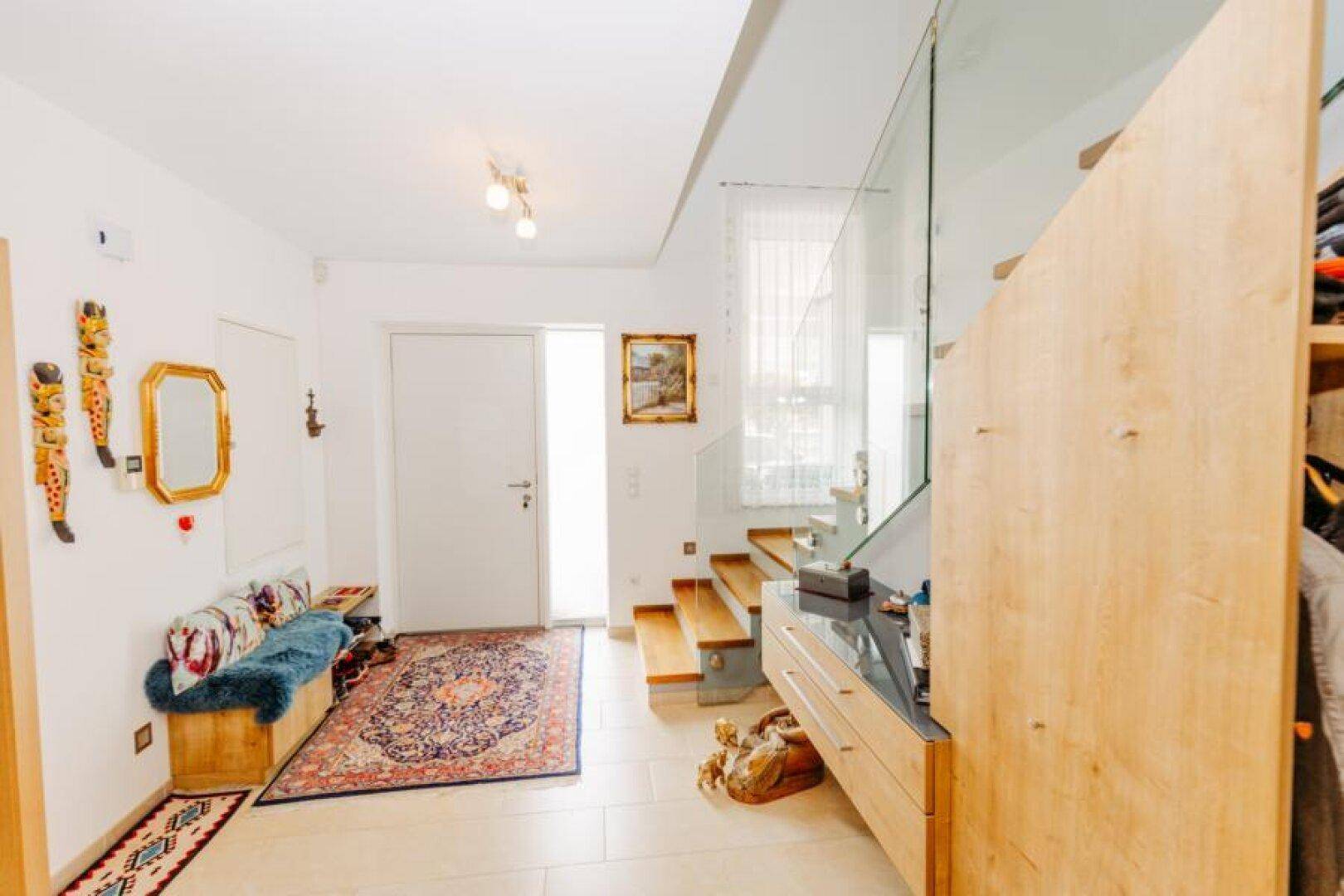 Heller Eingangsbereich mit modernen Fliesen, einer Holzbank und einer eleganten Glastreppe.