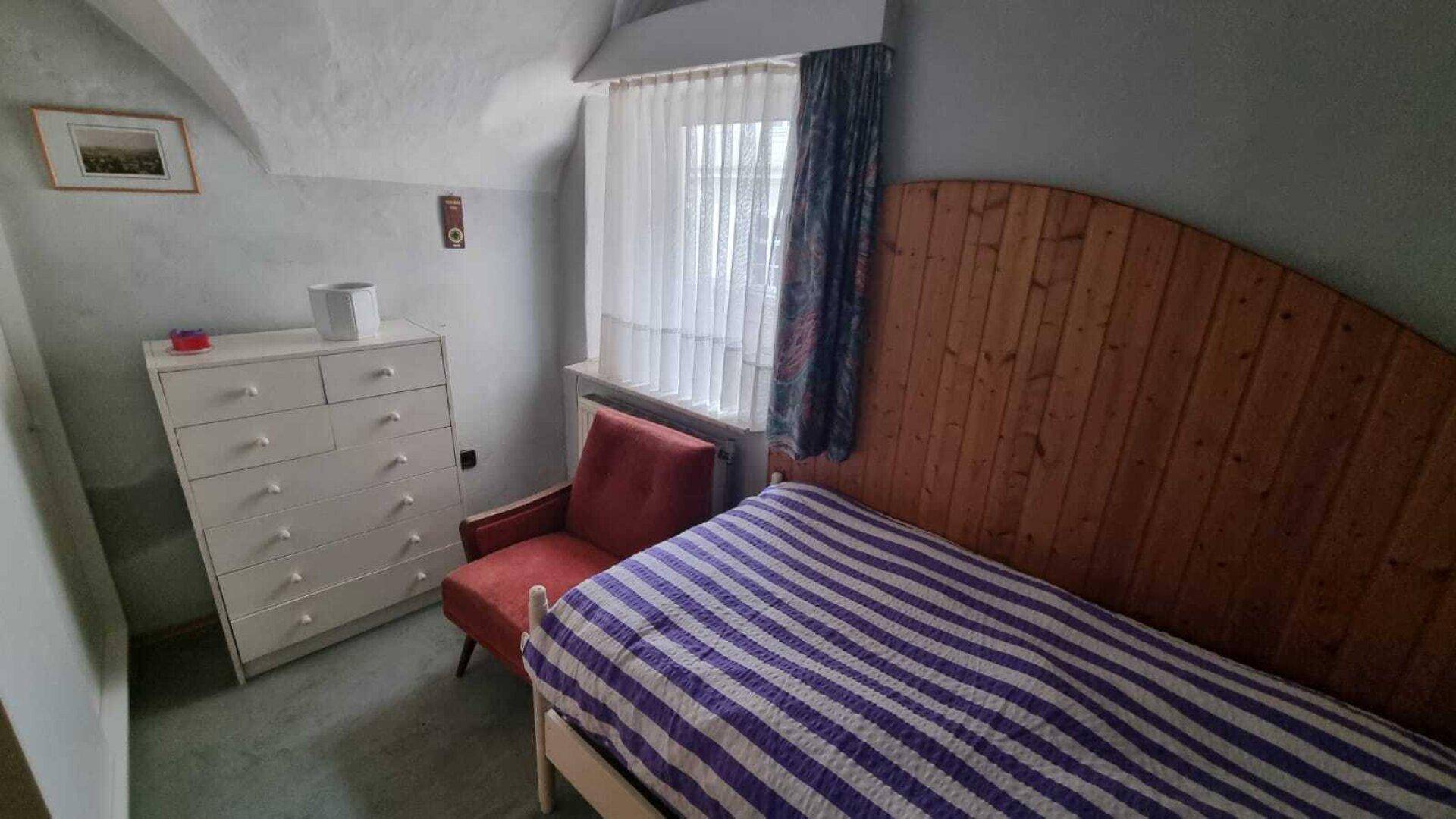 Helles Schlafzimmer mit Einzelbett, Kommode und einem Fenster mit Vorhängen.