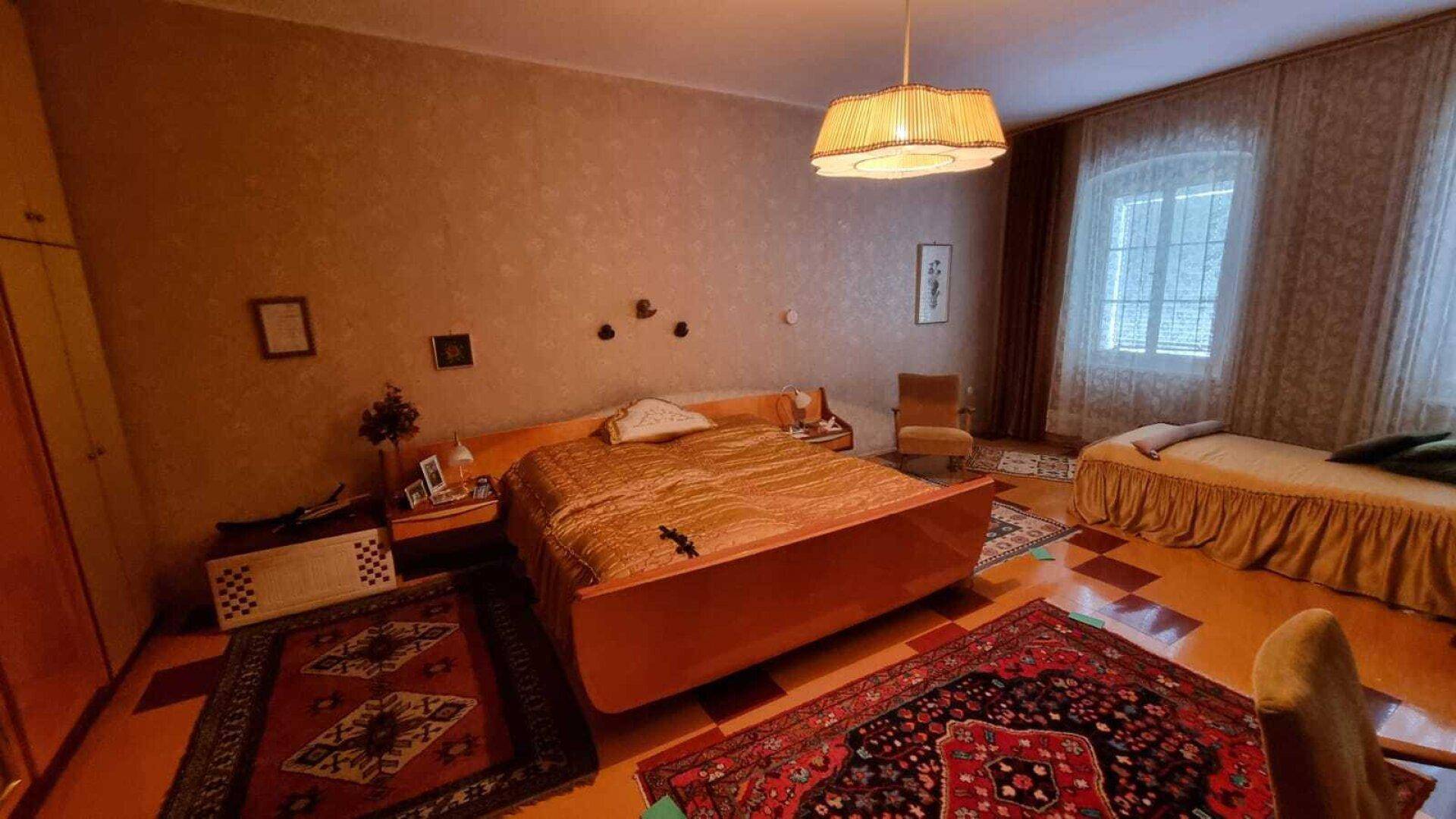 Schlafzimmer mit Doppelbett, Einzelbett, Teppichen und einem Fenster mit Vorhängen.