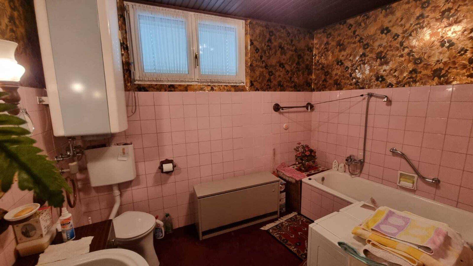 Badezimmer im Retro-Stil mit rosa Fliesen, Badewanne und einem Fenster mit floraler Tapete.