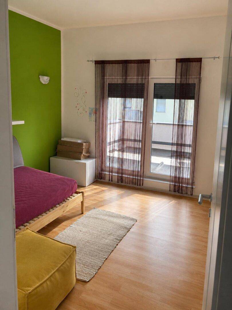 Schlafzimmer mit grün akzentuierter Wand, Holzboden und Zugang zum Balkon.