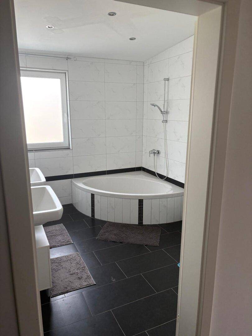 Badezimmer mit Eckbadewanne, Duschvorrichtung und dunklen Bodenfliesen.