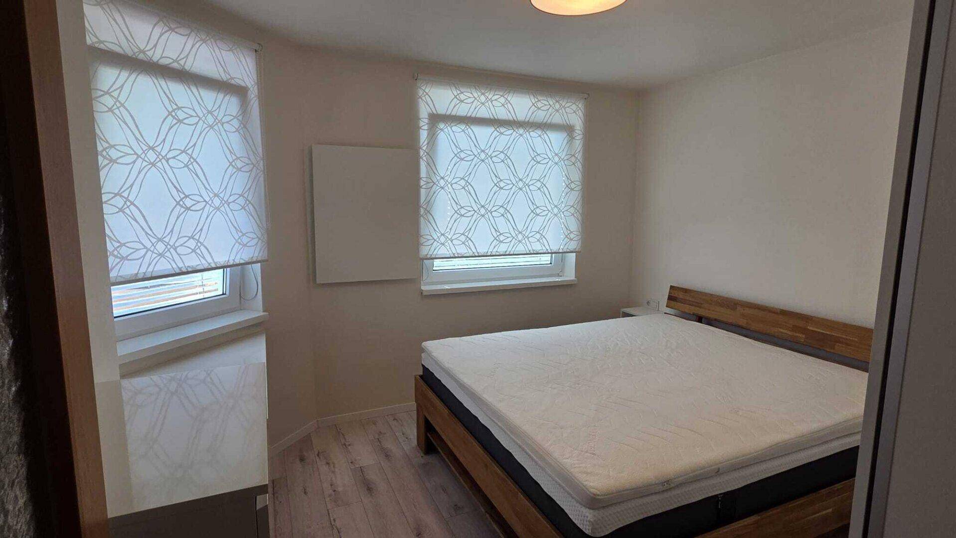 Schlafzimmer mit hellem Laminatboden, zwei Fenstern und einem einfachen Holzbett.