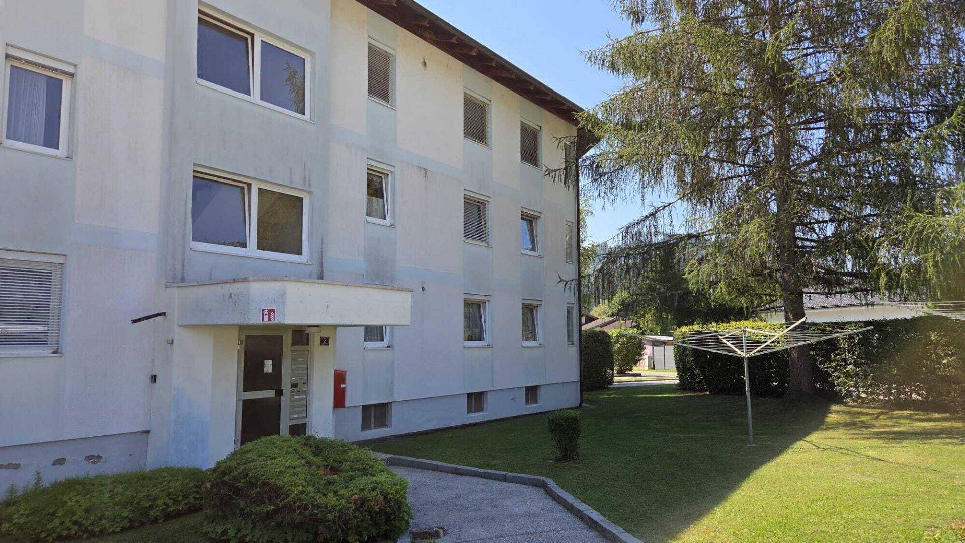 Eingangsbereich eines Mehrfamilienhauses mit gepflegter Grünfläche und Bäumen im Vordergrund.