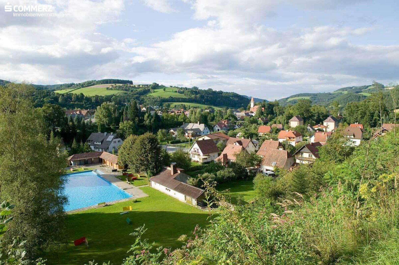 Panoramablick auf eine ländliche Siedlung mit Häusern, grünen Hügeln und einem Freibad.