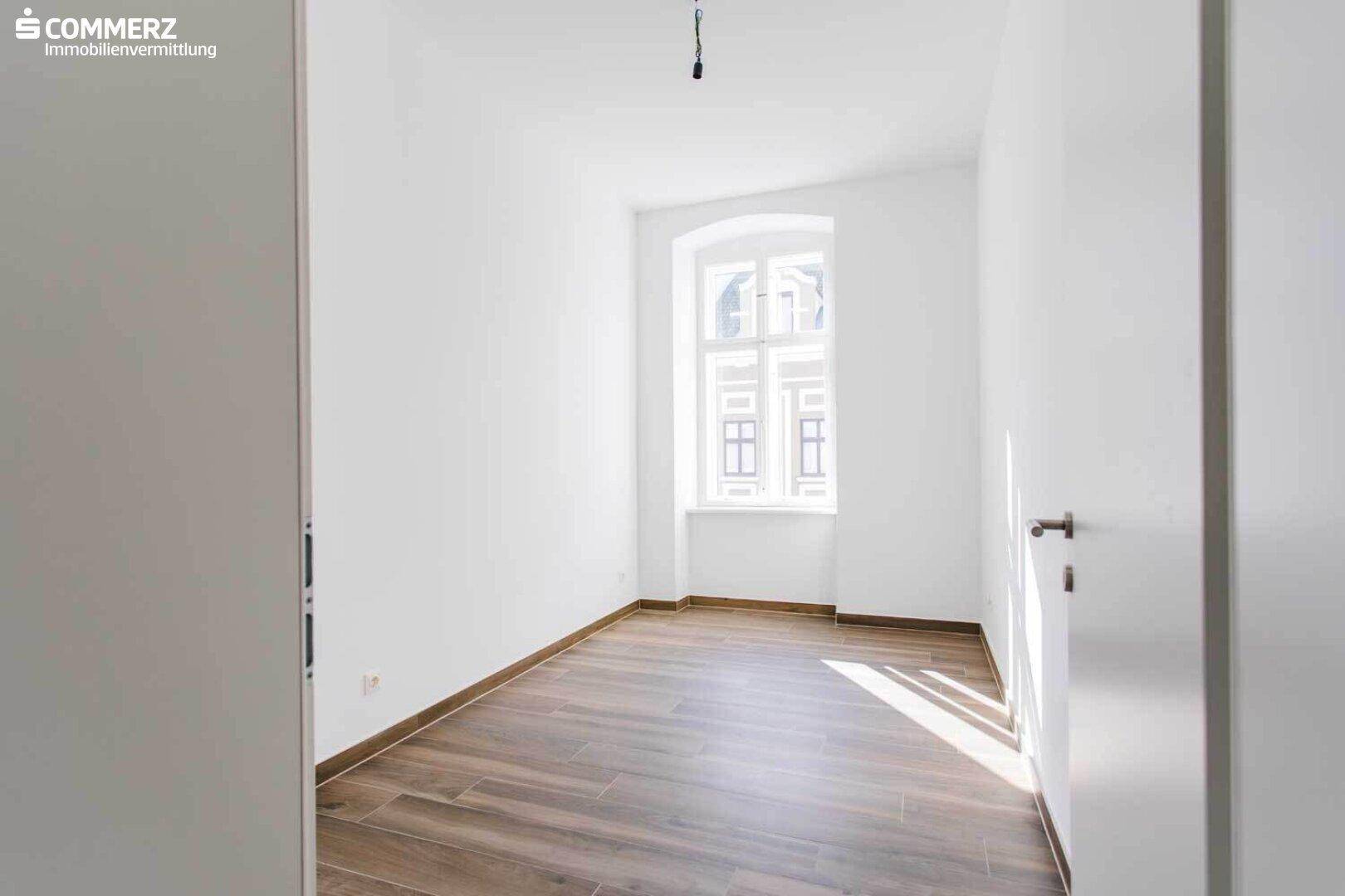 Helles Schlafzimmer mit Laminatboden und einem großen Fenster, das viel Tageslicht hereinlässt.
