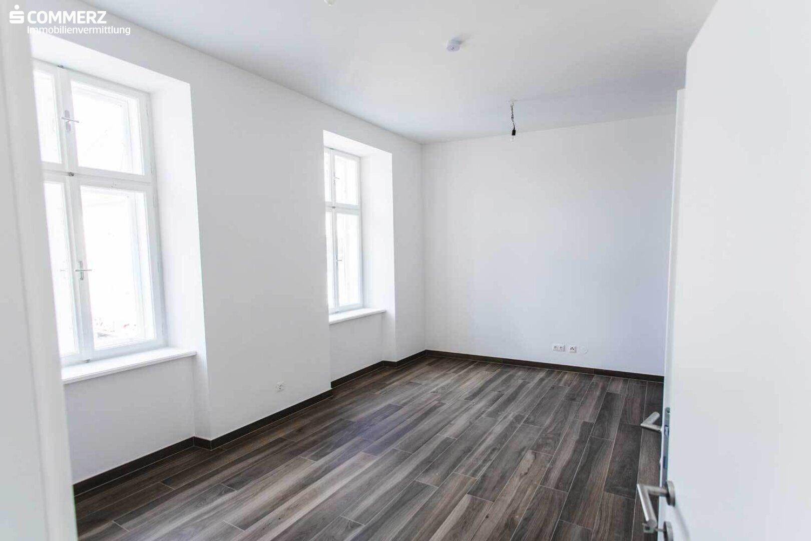 Unmöblierter Raum mit dunklem Laminatboden und hellen Wänden, durchflutet von natürlichem Licht.