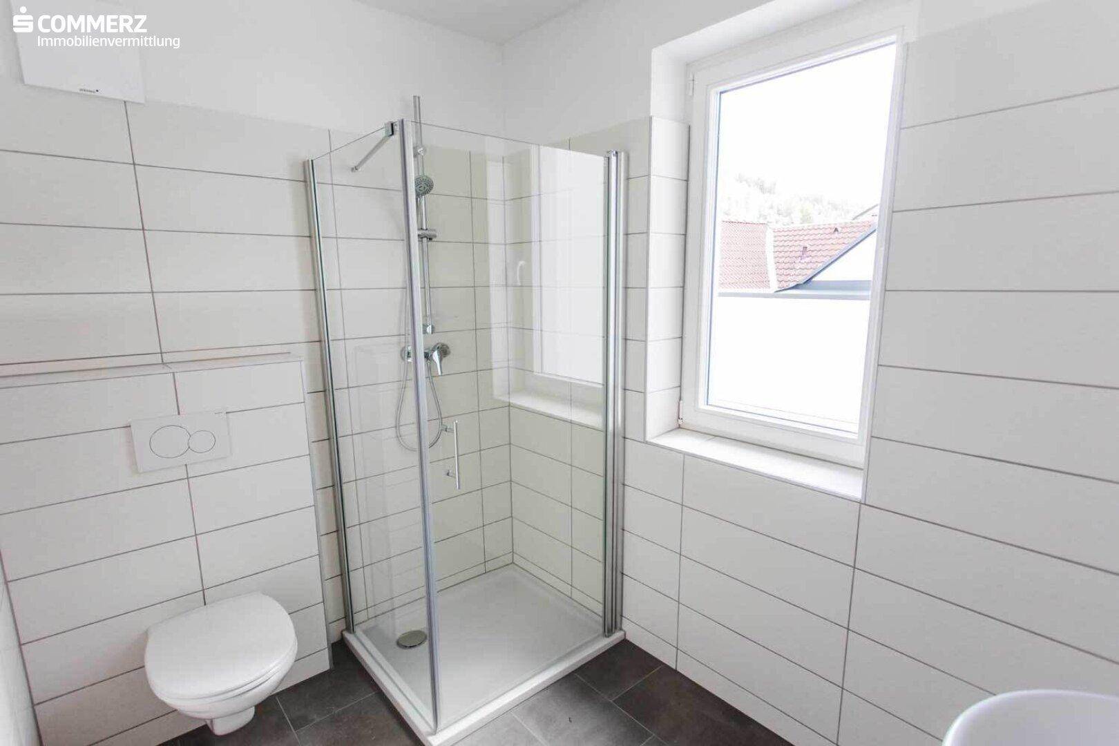 Modernes Badezimmer mit weißen Fliesen, dunklem Boden, WC und einer Duschkabine mit Fenster.