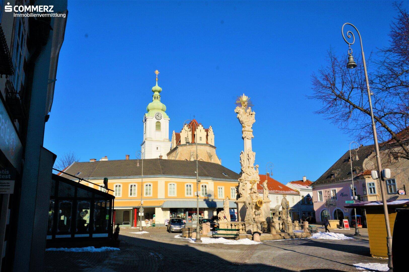 Historischer Stadtplatz mit Kirche, Denkmal und umliegenden Gebäuden unter klarem Winterhimmel.