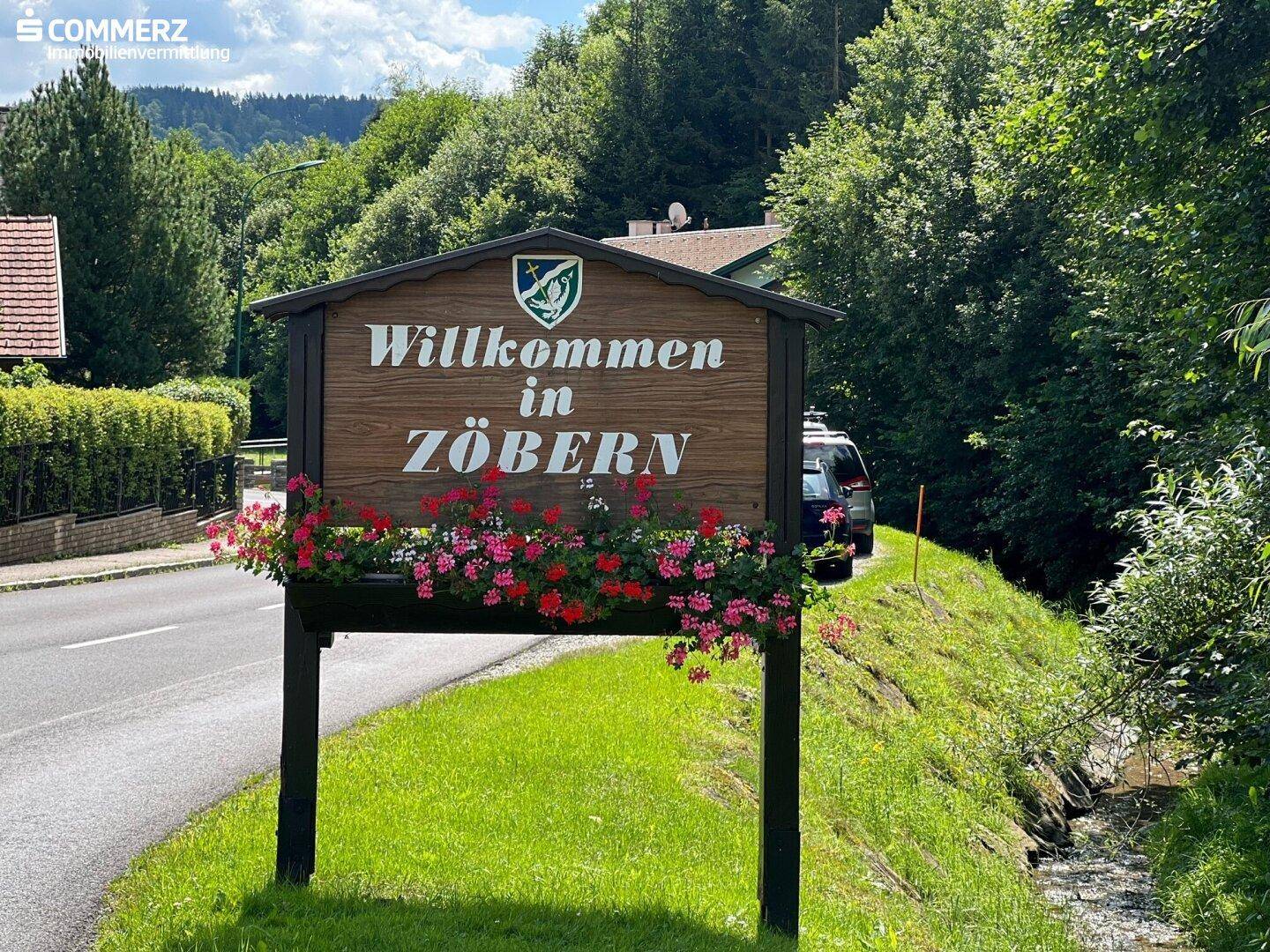 Willkommensschild in Zöbern mit Blumen, umgeben von grüner Landschaft und Bäumen am Straßenrand.