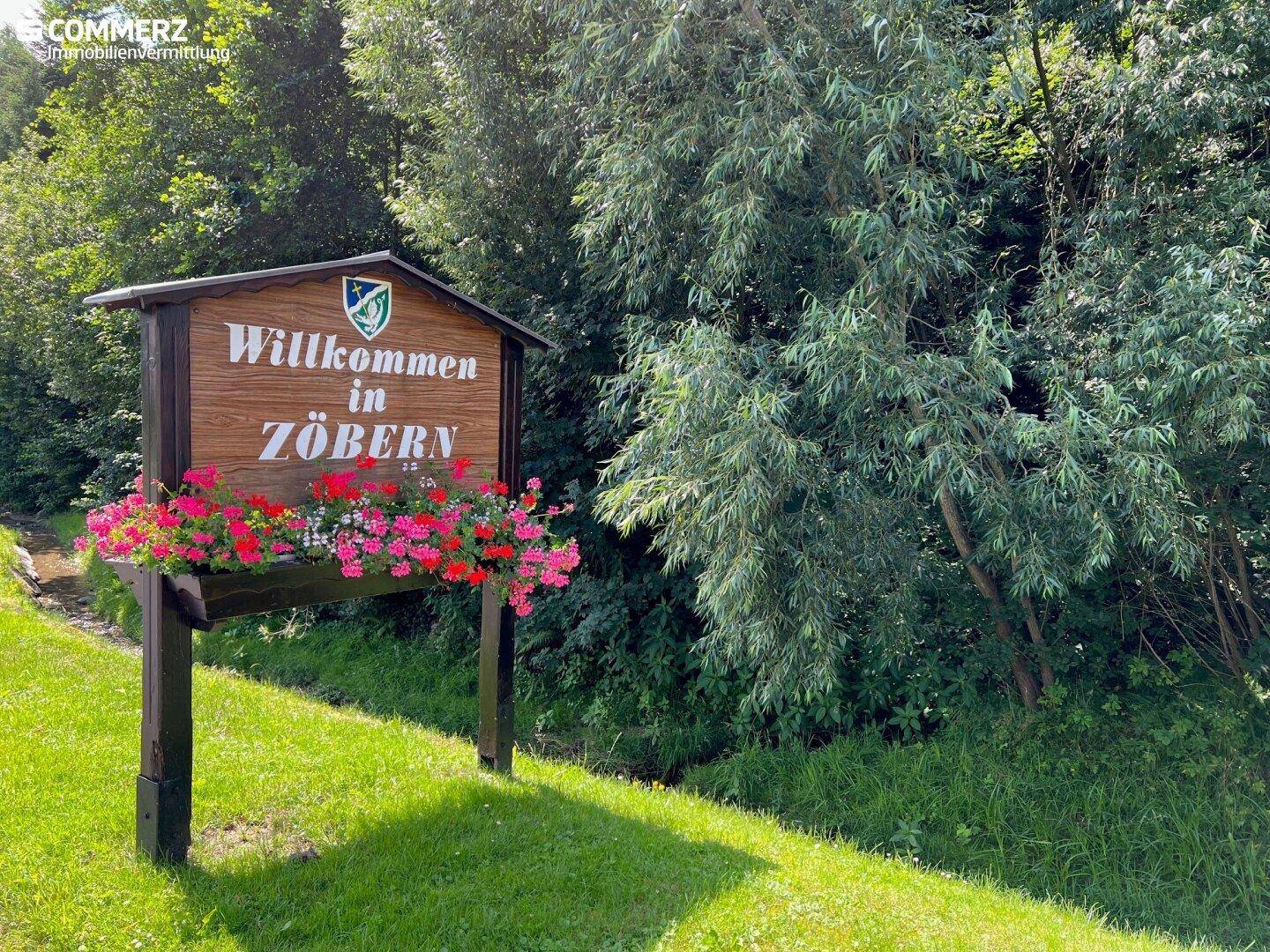 Willkommensschild in Zöbern mit roten Blumen, umgeben von dichtem Grün und Bäumen am Wegesrand.