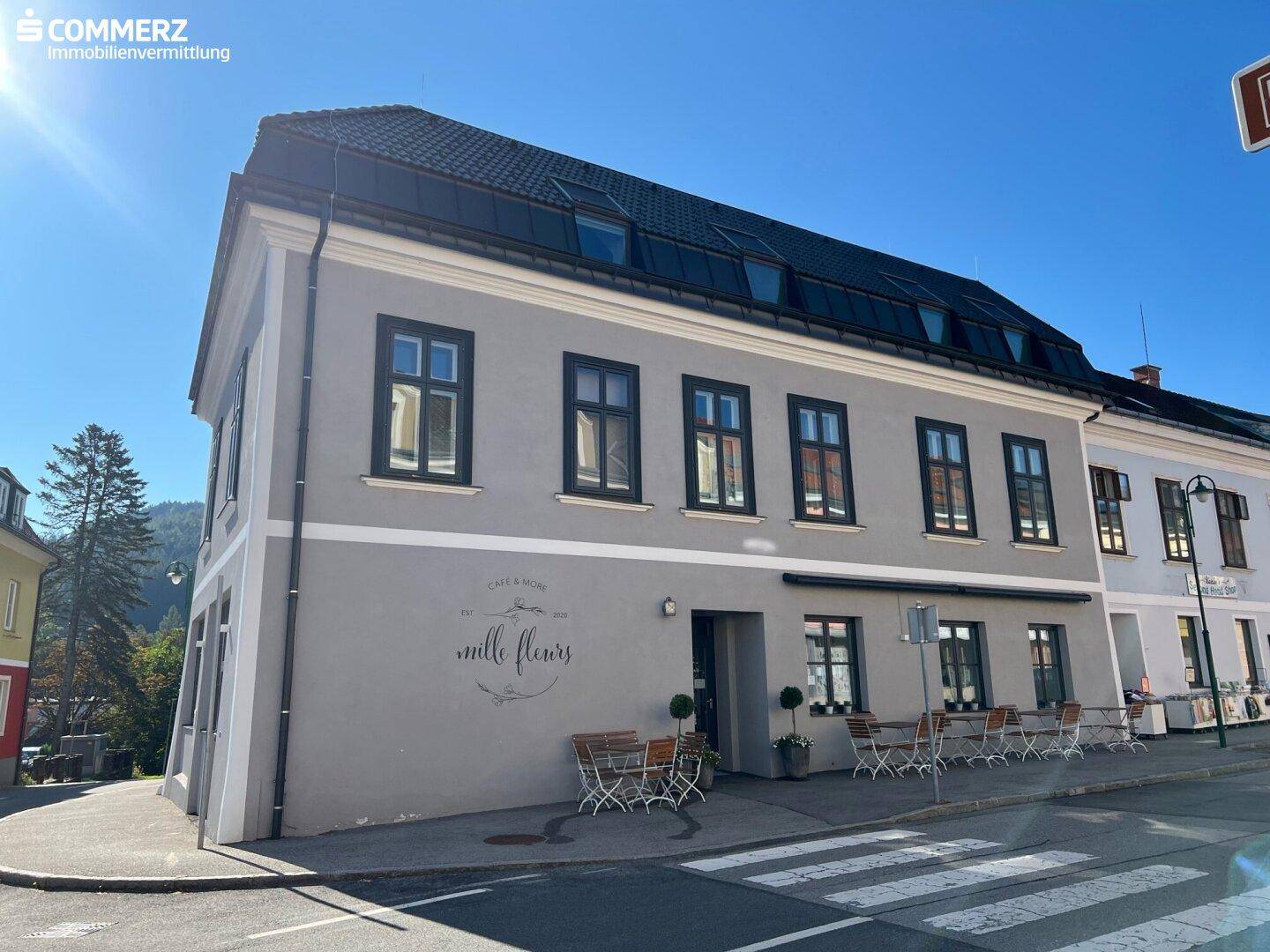 Modernisiertes Gebäude mit grauer Fassade, Mansarddach und Außensitzplätzen unter blauem Himmel.