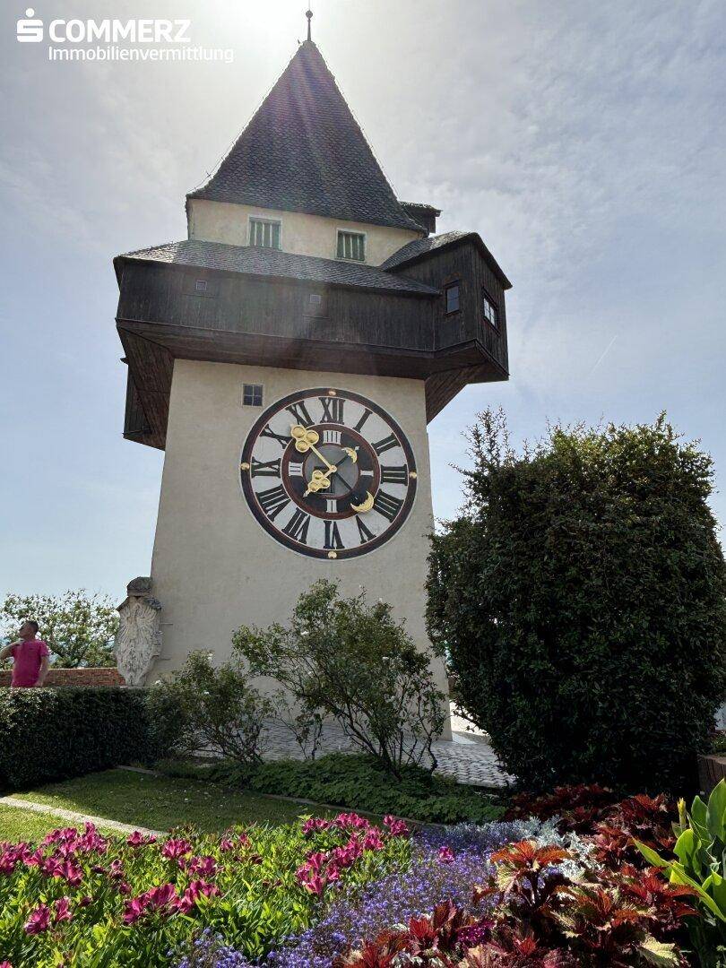 Der Grazer Uhrturm an einem sonnigen Tag, umgeben von bunten Blumenbeeten.