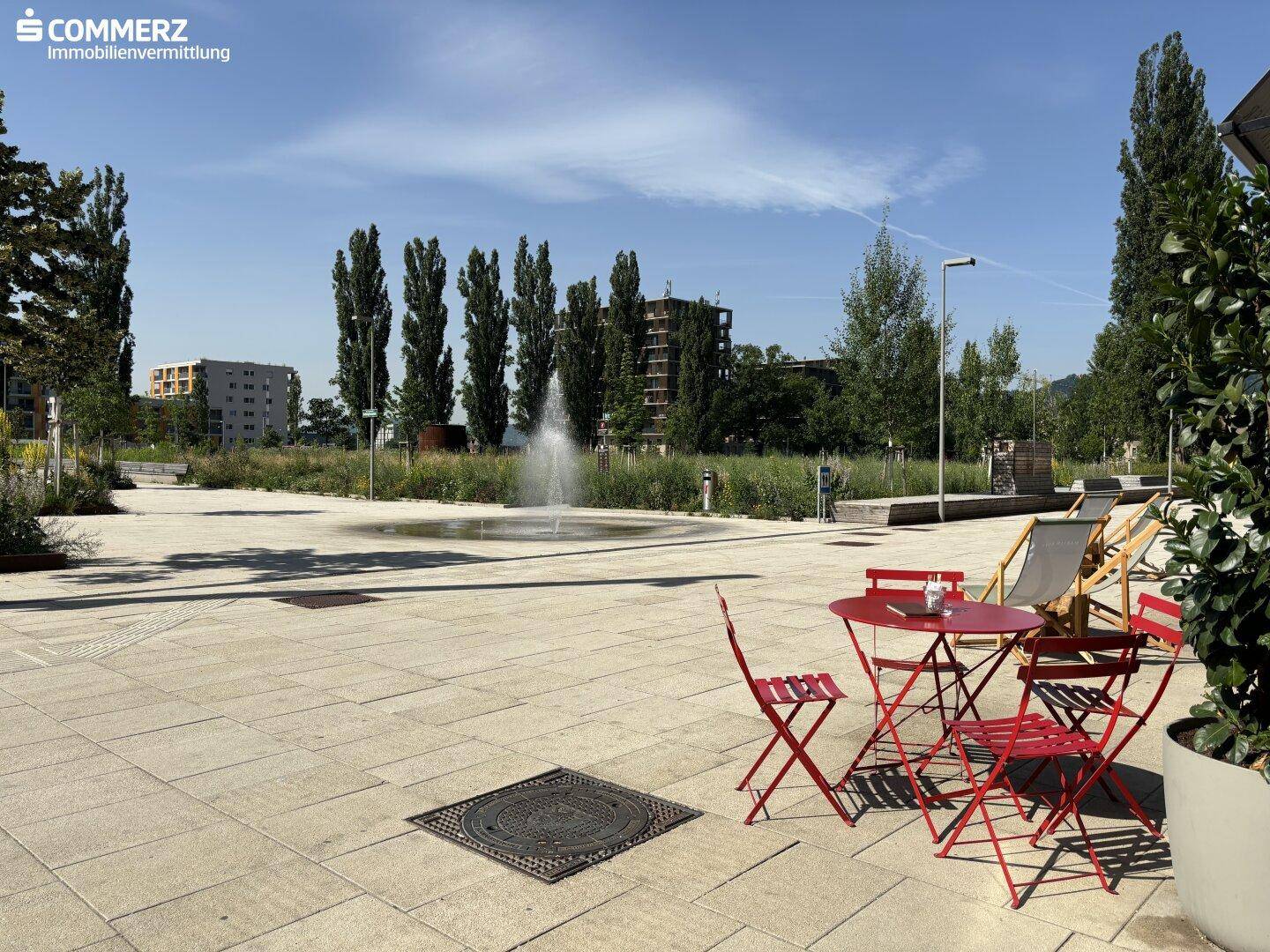 Öffentlicher Platz mit Sitzgelegenheiten, einem Springbrunnen und Bäumen unter blauem Himmel.