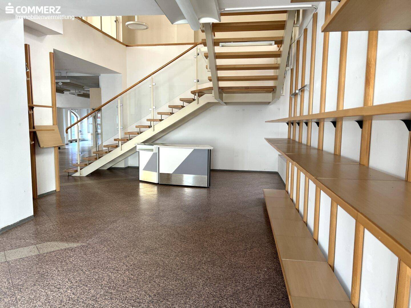 Heller Innenraum mit moderner Holztreppe, Glasgeländer und Regalsystemen, ideal für Verkaufsflächen.