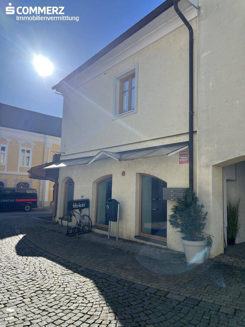 Fassade eines historischen Gebäudes mit Schaufenstern und Kopfsteinpflaster im Sonnenlicht.