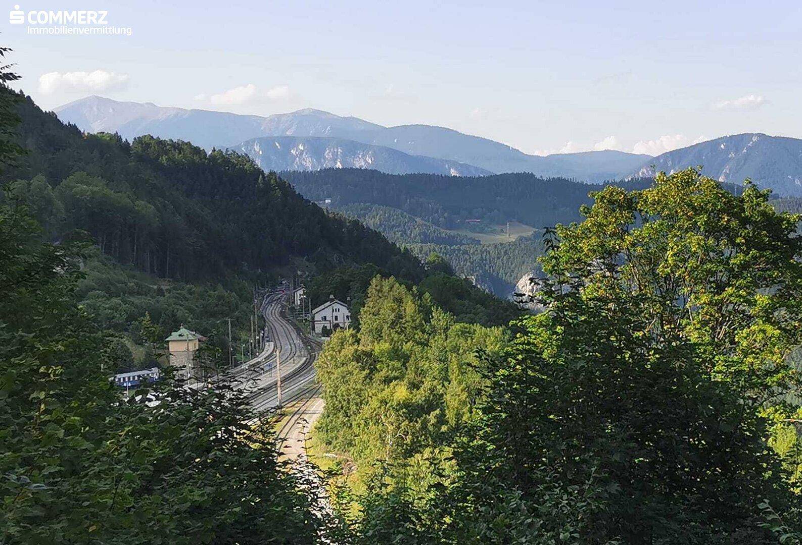Panoramablick auf eine grüne Berglandschaft mit einer Bahnstrecke und vereinzelten Gebäuden.