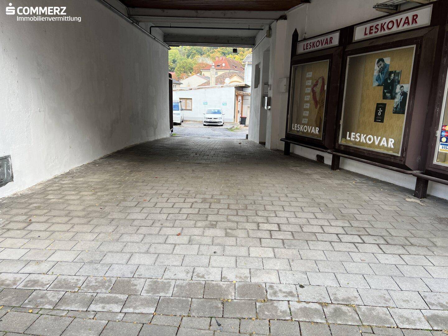 Gepflasterte Zufahrt zum Innenhof, flankiert von Gebäudeteilen und Schaufenstern.