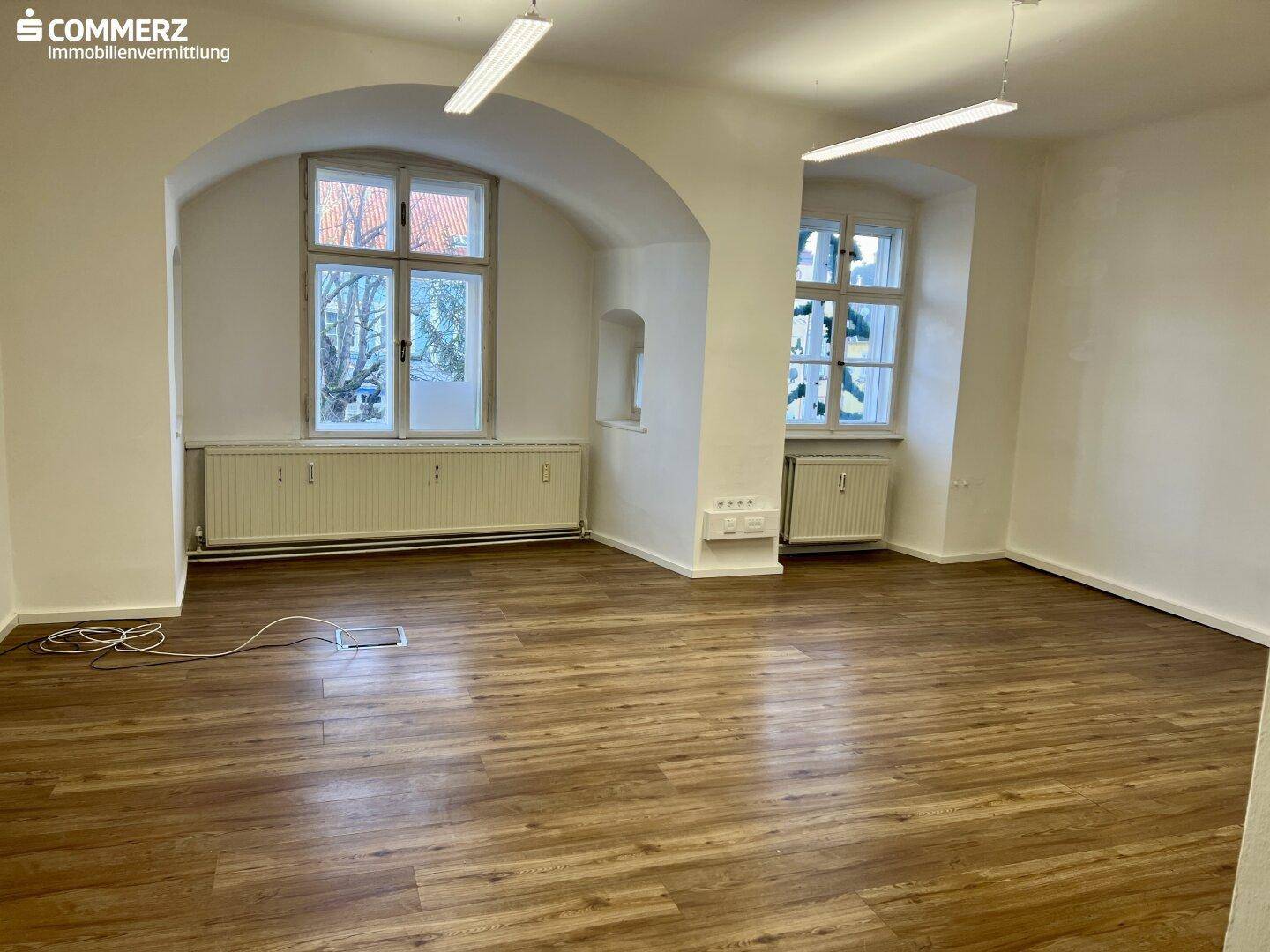 Geräumiges Büro mit Laminatboden, hohen Decken und zwei großen Bogenfenstern für viel Tageslicht.