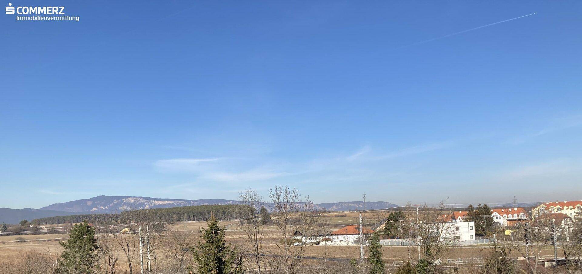 Weitläufiger Blick über Felder und Hügel mit Bergen am Horizont unter strahlend blauem Himmel.