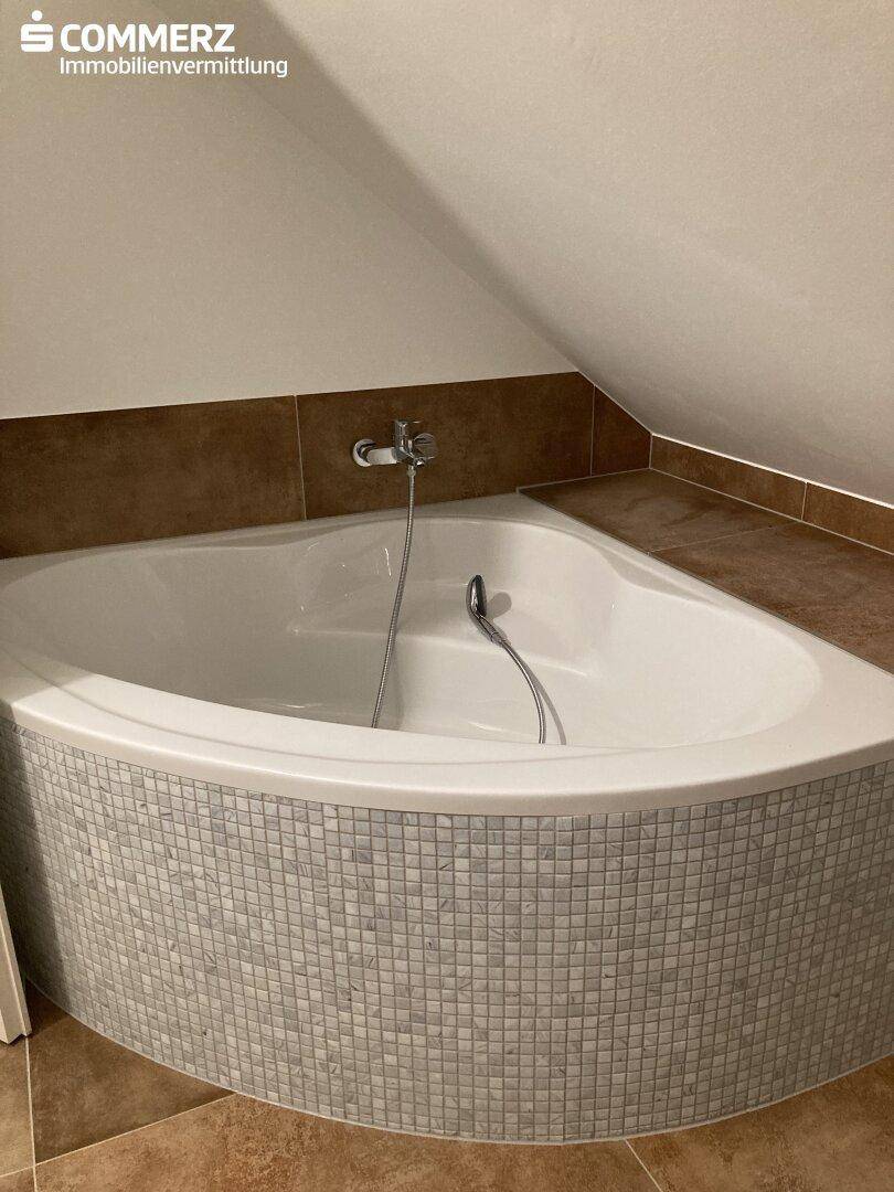 Badezimmer im Dachgeschoss mit Eckbadewanne und braunen Fliesen an Wänden und Boden.