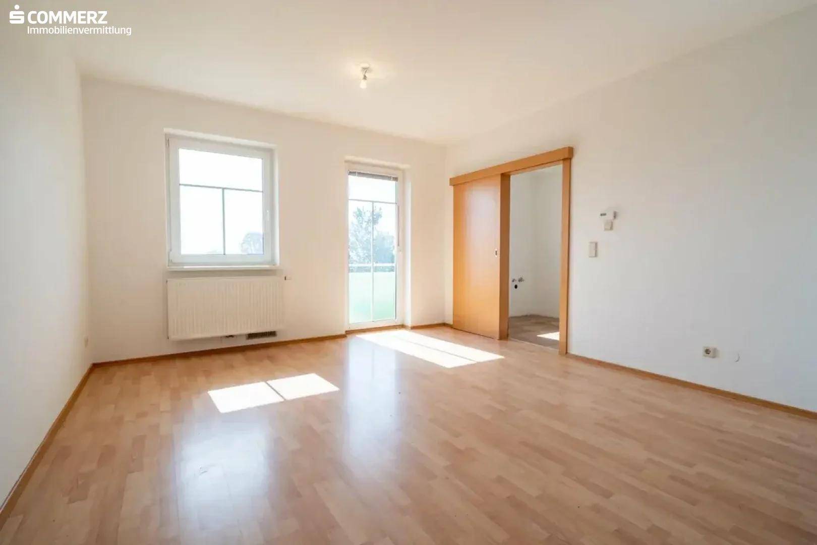 Helles, leeres Wohnzimmer mit Laminatboden, großem Fenster und Zugang zum Balkon.