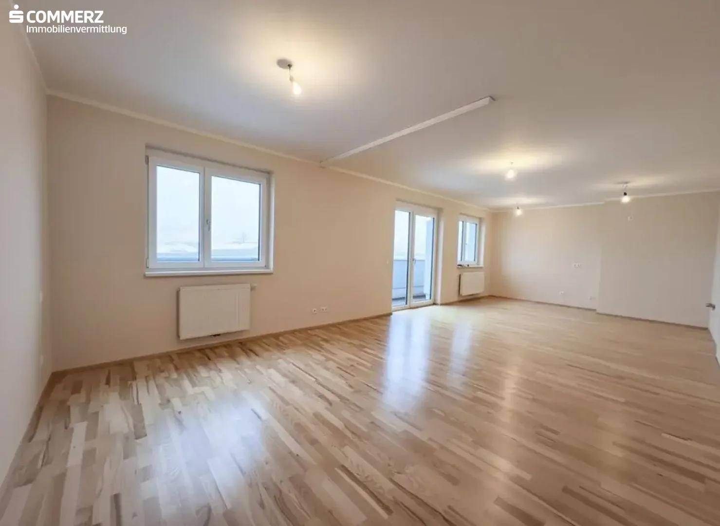 Geräumiges, leeres Wohnzimmer mit hellem Parkettboden und Zugang zum Balkon.