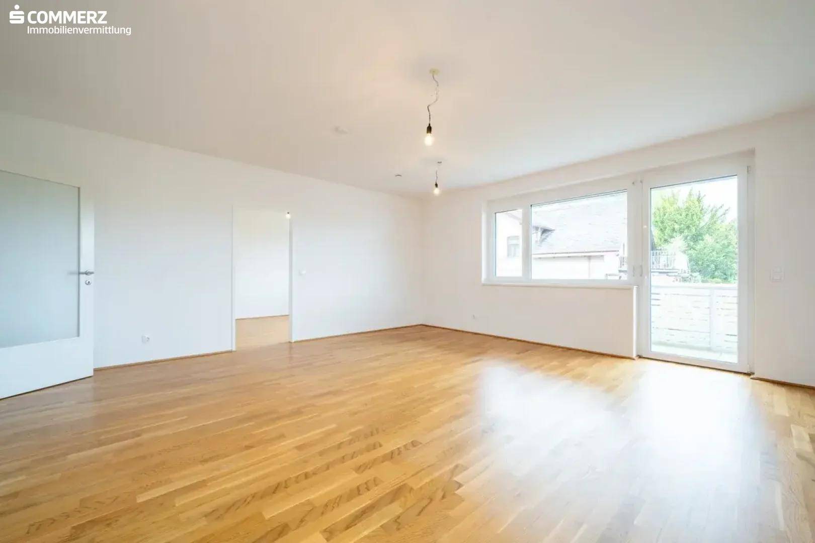 Helles, leeres Zimmer mit Holzboden und Zugang zu einem Balkon oder einer Terrasse.