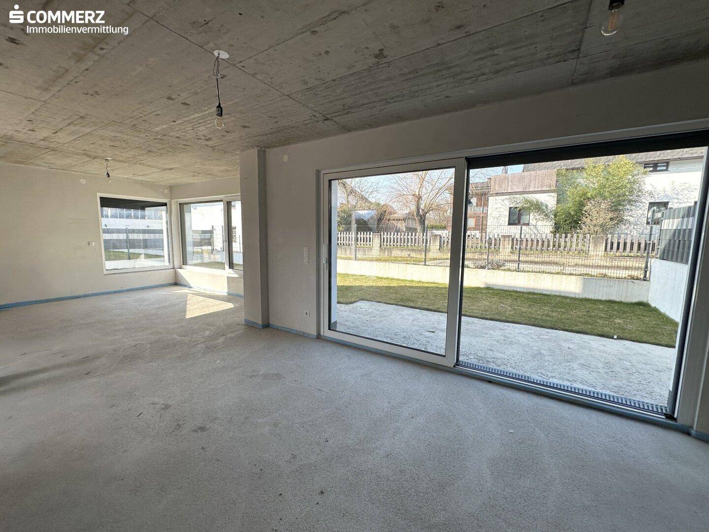 Unfertiger Innenraum mit Betonboden und großen Fenstern zum Gartenbereich.