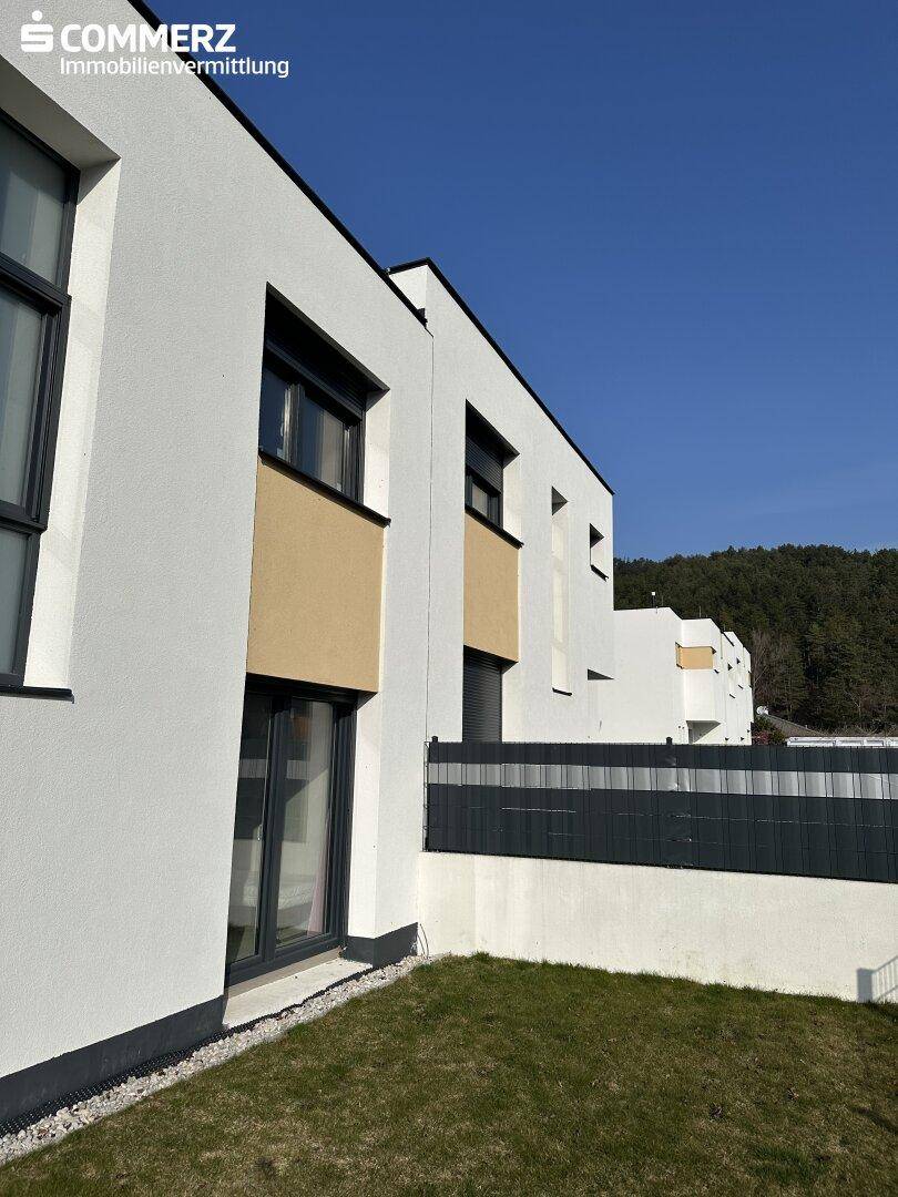 Gepflegte Rasenfläche neben einem modernen Reihenhaus mit dunklen Fensterrahmen und Bergblick.
