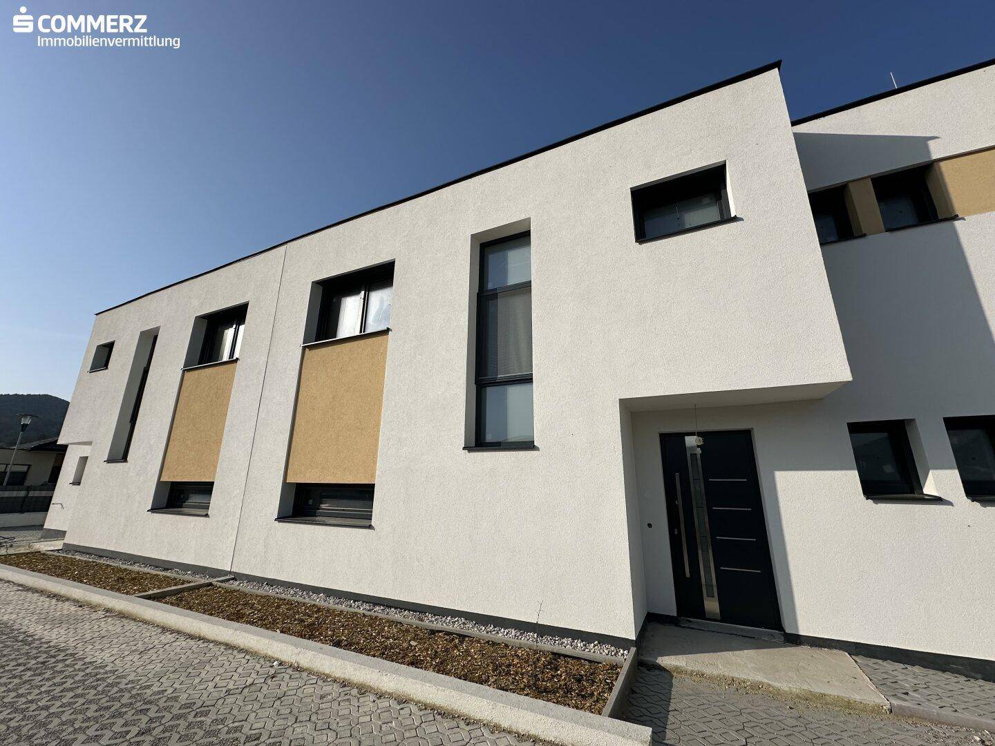 Frontansicht eines modernen Reihenhauses mit klaren Linien und großen Fenstern unter blauem Himmel.