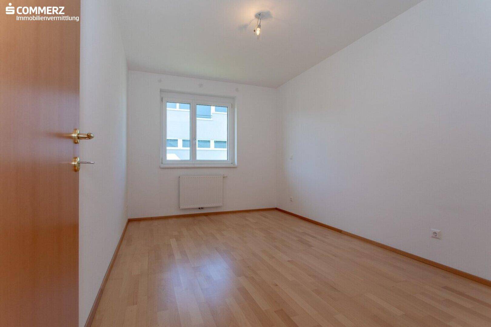 Ruhiges Schlafzimmer mit hellem Parkettboden und einem Fenster mit Blick auf das Nachbargebäude.