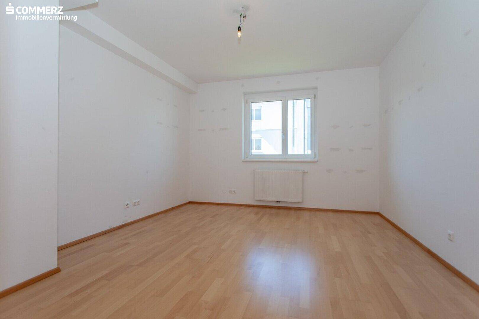 Leeres Zimmer mit Parkettboden und einem Fenster, das viel Tageslicht hereinlässt.