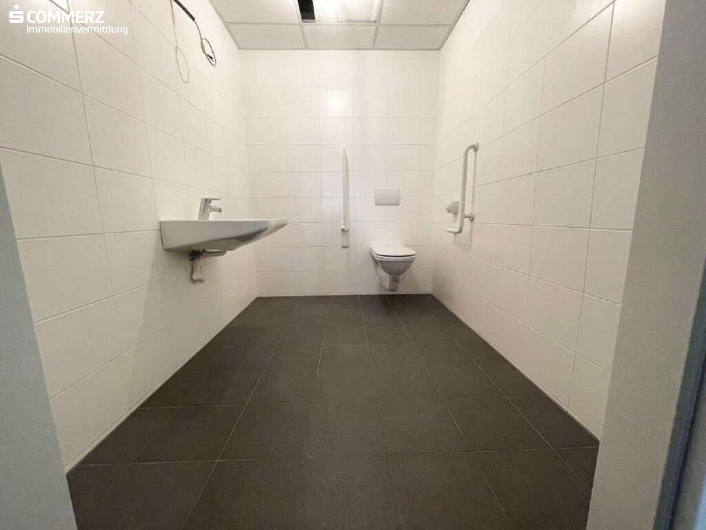 Modernes, barrierefreies Badezimmer mit weißen Fliesen, dunklem Boden und Haltegriffen an der Wand.