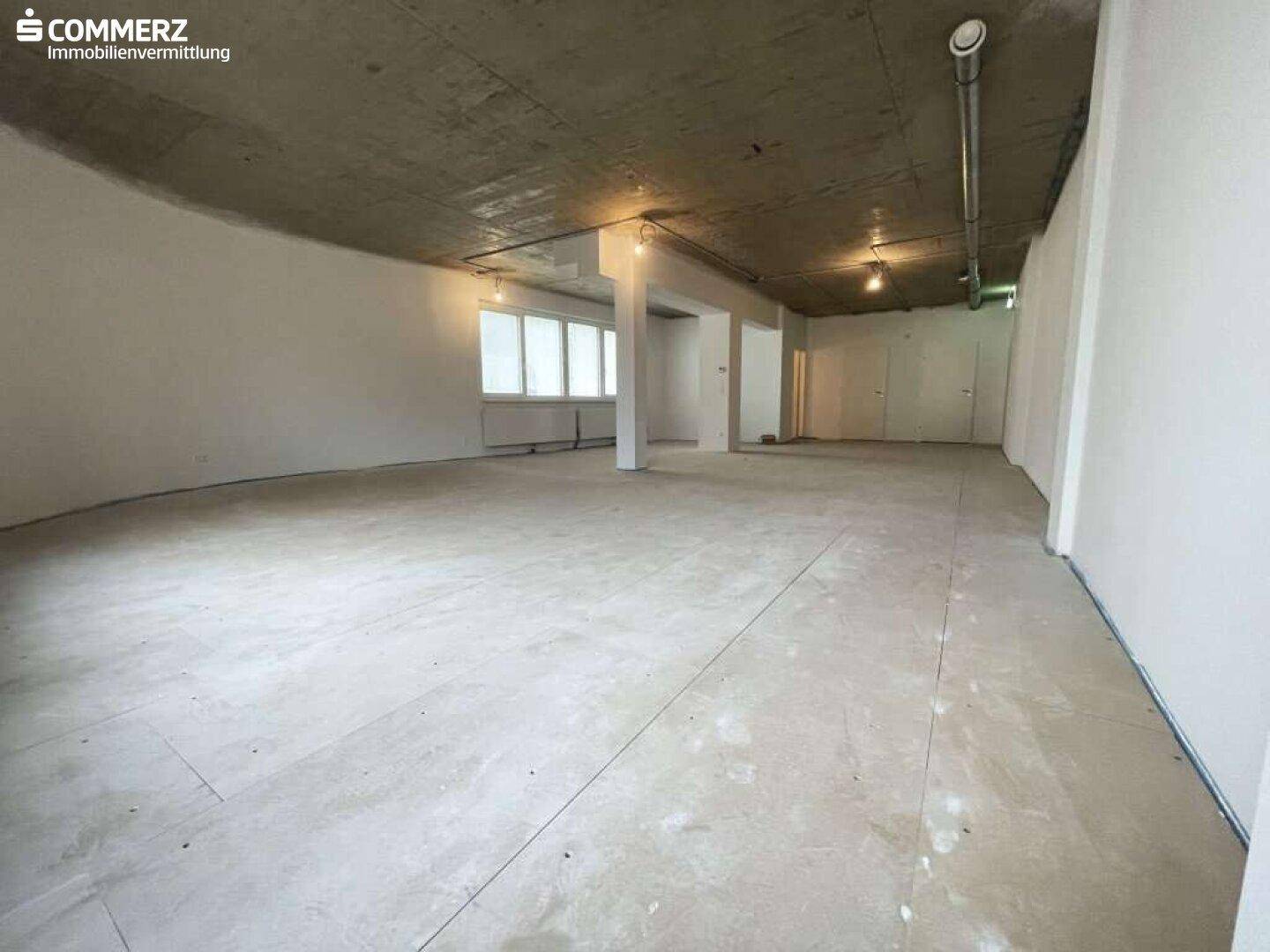 Weitläufiger, leerer Raum mit Betonboden und sichtbaren Deckeninstallationen, bereit für den Innenausbau.