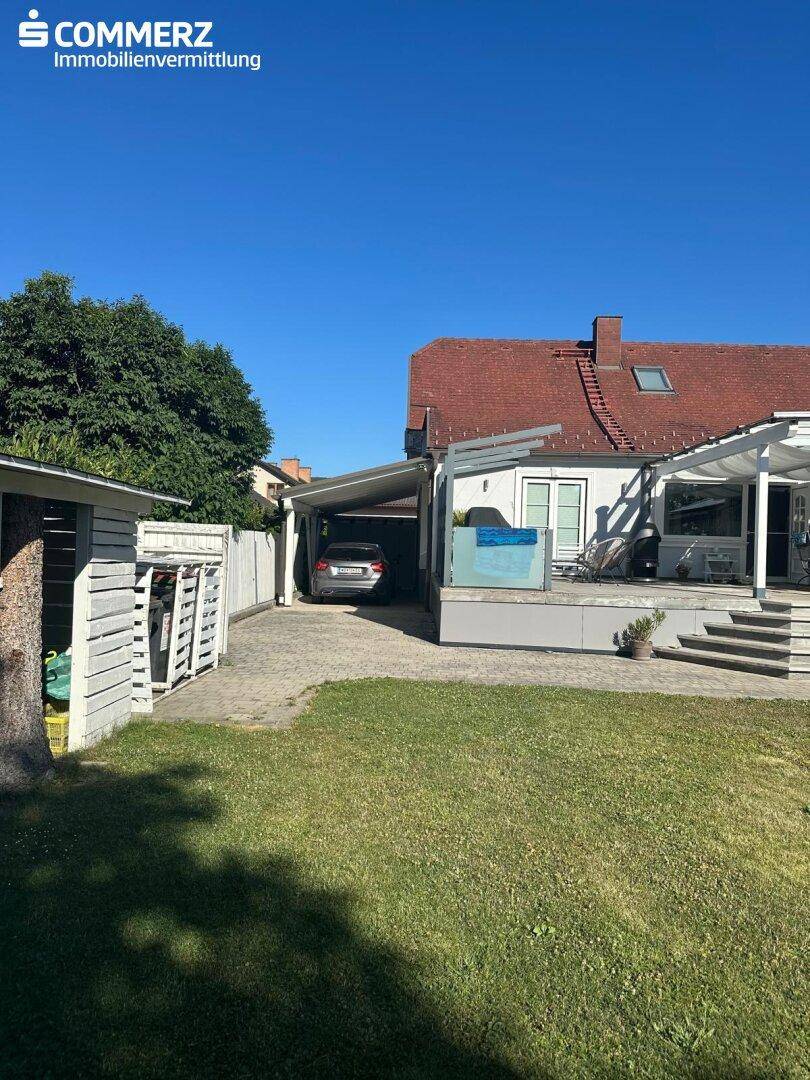 Seitenansicht des Hauses mit Carport, Zufahrt und einem kleinen Gartenhaus im Vordergrund.