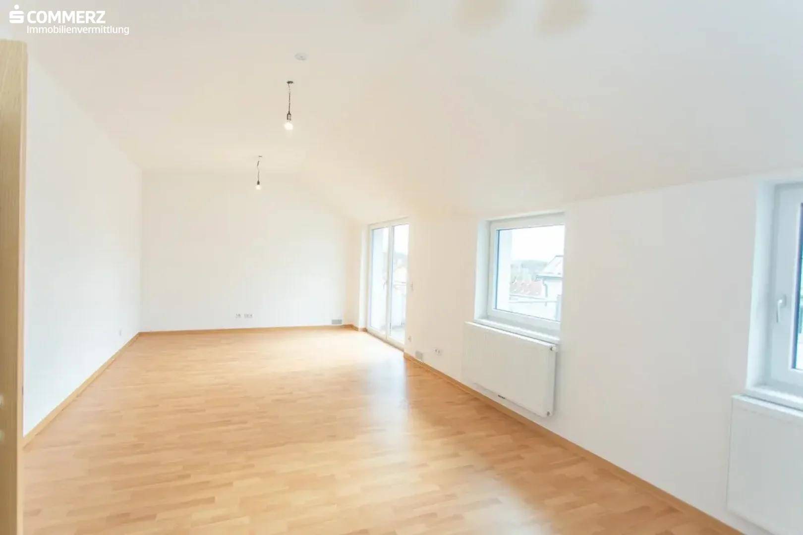 Helles, leeres Wohnzimmer mit Holzboden, großen Fenstern und Zugang zum Balkon.