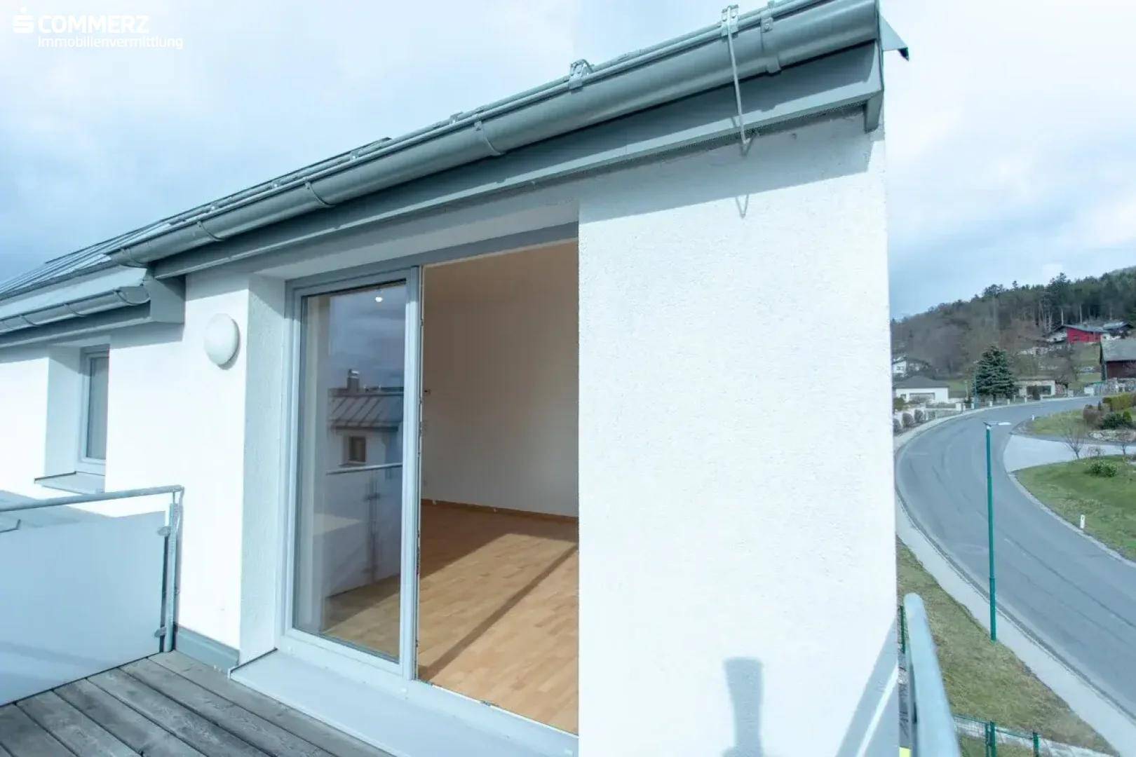 Balkon mit Holzboden und Schiebetür, bietet einen Blick auf die Straße und grüne Hügel.