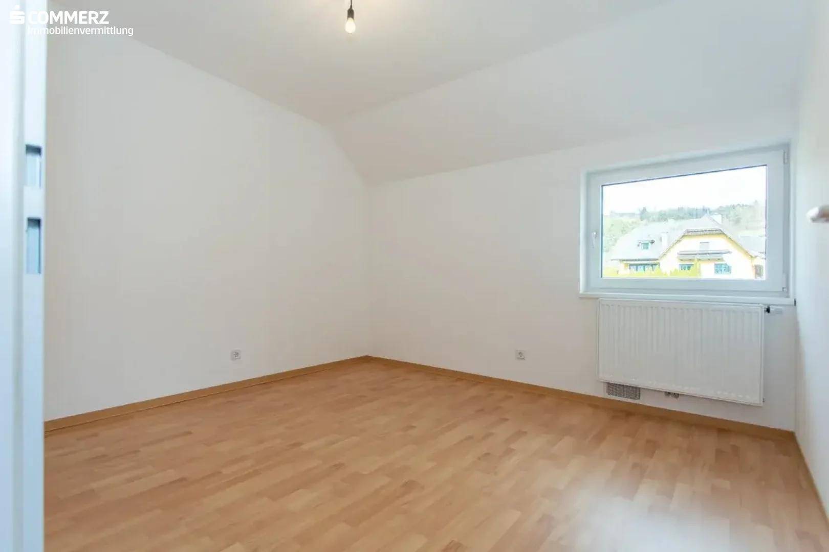 Helles Schlafzimmer mit Holzboden, weiß gestrichenen Wänden und Fensterblick ins Grüne.