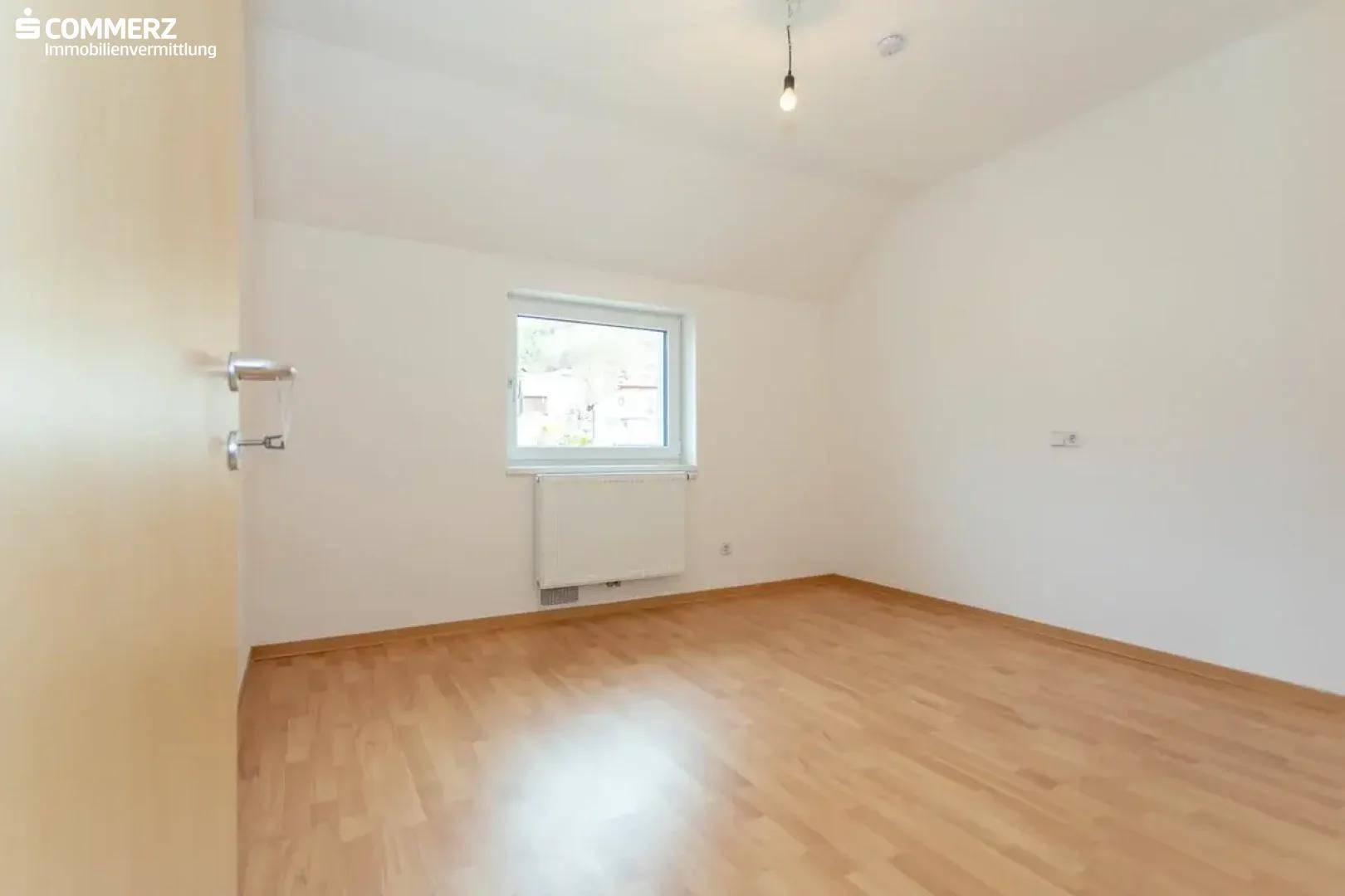 Geräumiges Schlafzimmer mit Holzboden, weiß gestrichenen Wänden und einem großen Fenster.