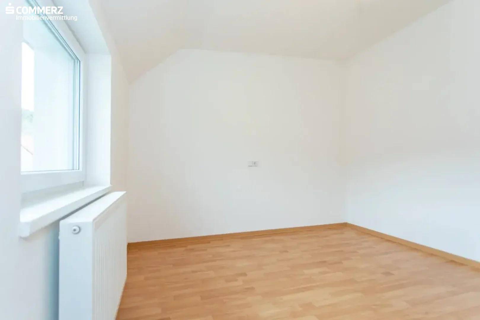 Schlafzimmer mit Holzboden, weiß gestrichenen Wänden und einem Fenster, das viel Licht hereinlässt.