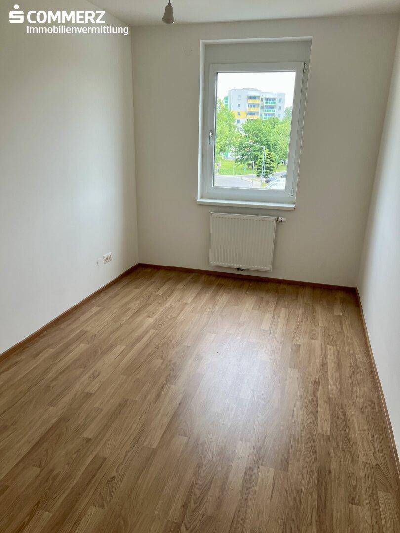 Leeres Zimmer mit Laminatboden und einem Fenster, das viel Tageslicht hereinlässt.