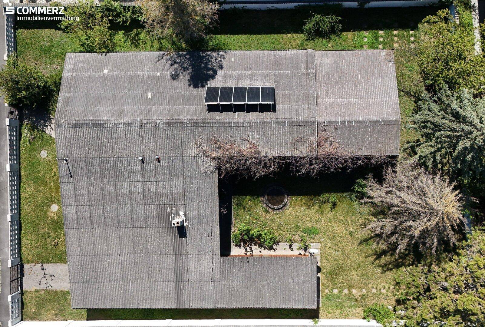 Luftaufnahme eines Gebäudes mit Wellblechdach, Solarpaneelen und umliegendem Gartenbereich mit Bäumen.