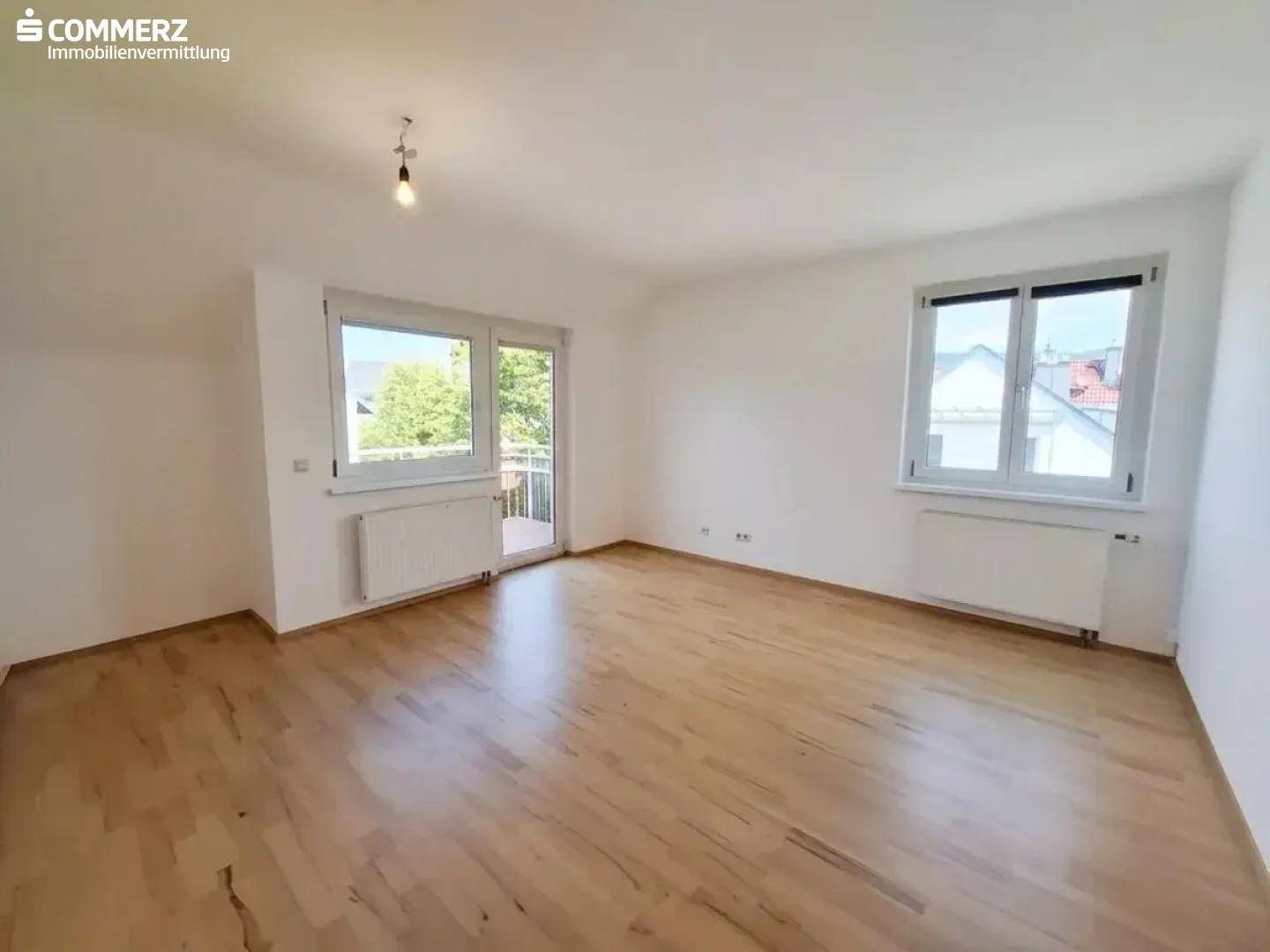 Helles, leeres Zimmer mit Parkettboden, großen Fenstern und Zugang zum Balkon.
