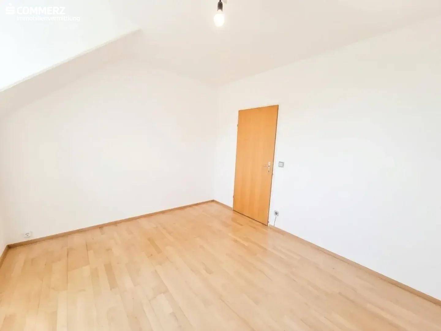 Helles Zimmer im Dachgeschoss mit Parkettboden, Dachschräge und Holztür.