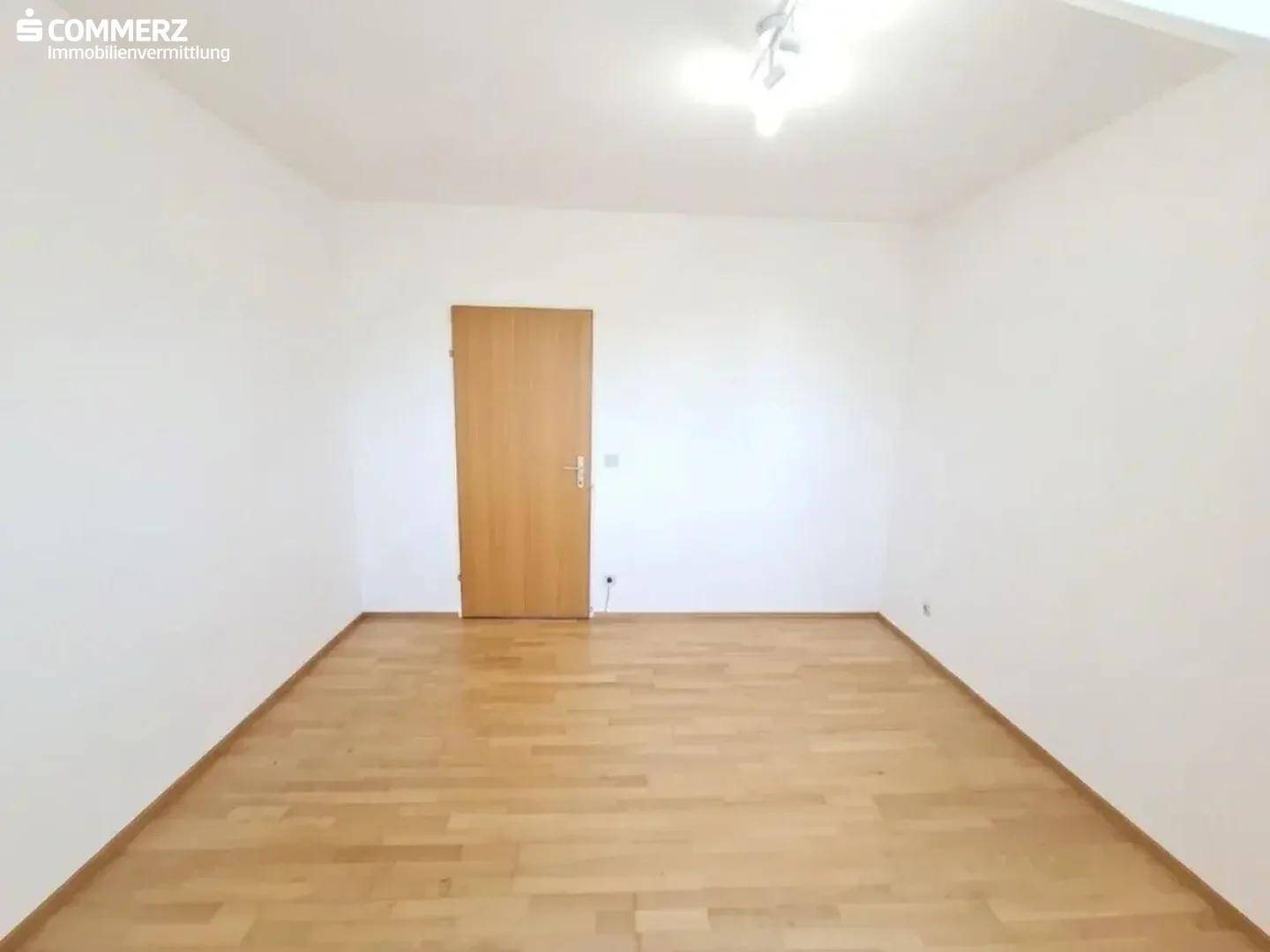 Geräumiges, helles Zimmer mit Parkettboden, weiß gestrichenen Wänden und Holztür.
