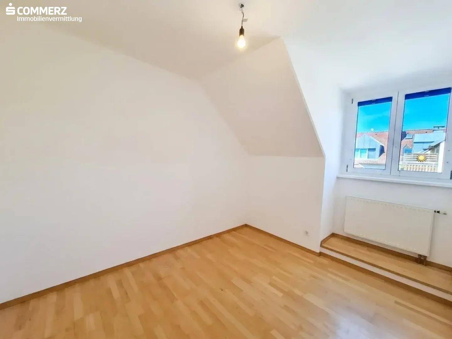 Helles Zimmer im Dachgeschoss mit Parkettboden, Dachschräge und Fenster mit Ausblick.