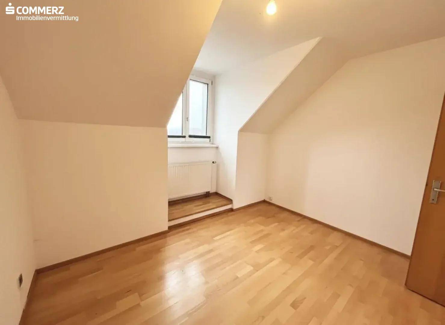 Helles Zimmer im Dachgeschoss mit Parkettboden, Dachschräge und Fenster.