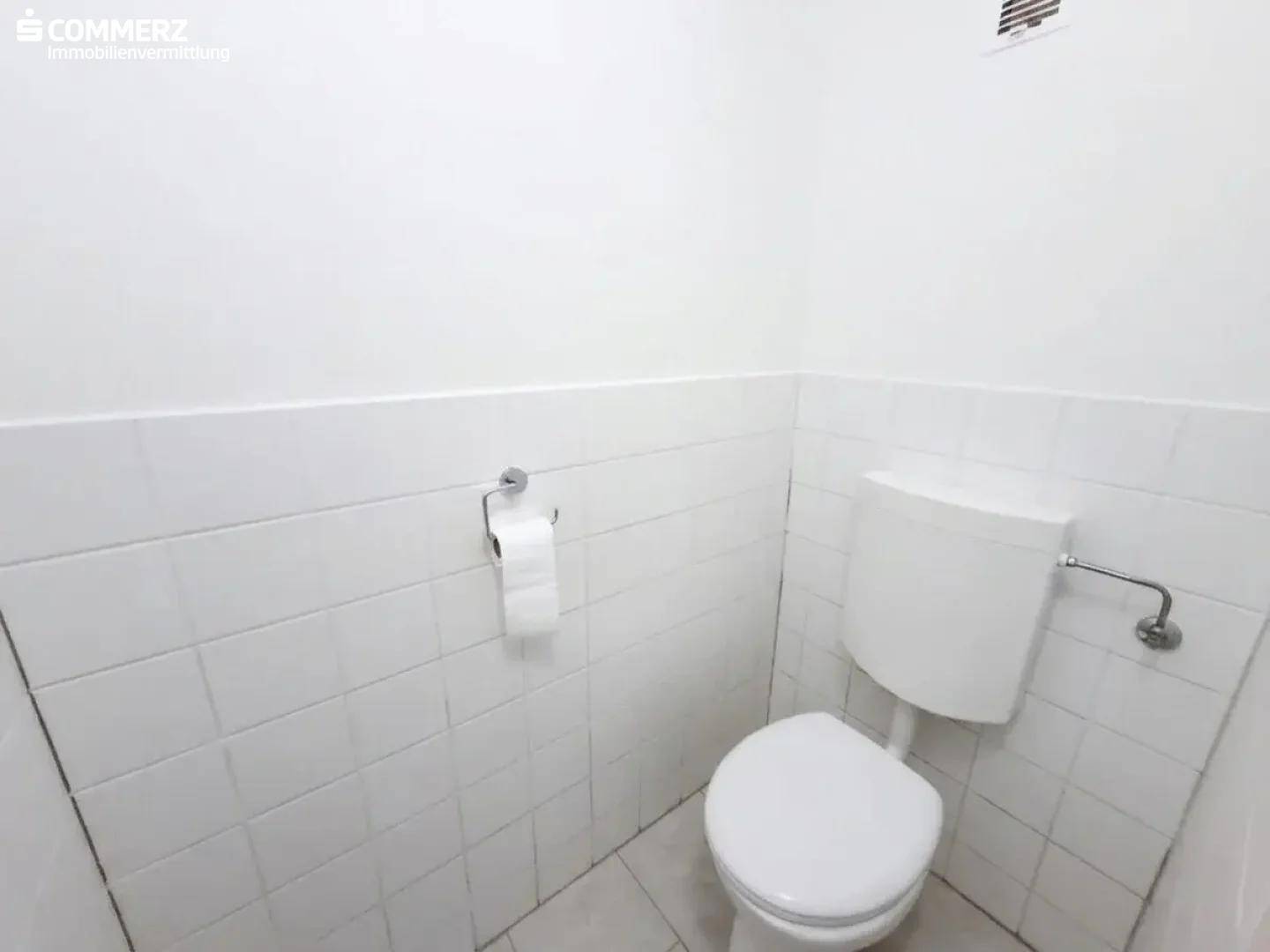 Separates WC mit weiß gefliesten Wänden, Toilette und Lüftungsgitter.